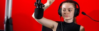 Female Multimedia Journalism student operating the control deck in the radio studio. They are wearing headphones and have their right hand raised, signalling to begin recording. They are photographed against a red background