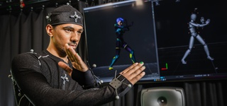 Male student using the motion capture facilities in our Screen School. They are wearing a special suit with sensors that records their movements. They have their hands crossed forming an X-shape