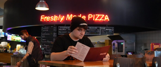 A student examining the menu in the Light cinema pizza restaurant, a neon sign in the background reading FRESHLY MADE PIZZA