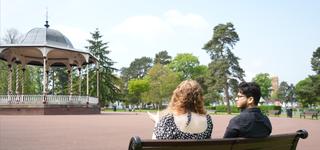 Two students in a park with many different trees, sitting on a bench which faces a sheltered area