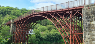 A fenced iron bridge crossing a body of water, comprising an ornate arch stylised with chain shapes, a house visible between the trees on the opposite side of the bridge