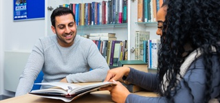 Photograph of two students sitting together in a library, sharing a book across a table.