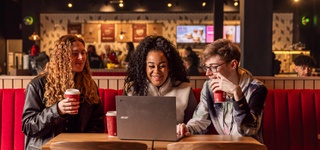 Three students sitting at a table in the on-campus coffee shop, looking at a single laptop between them