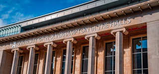 Photograph of the University of Wolverhampton at The Halls venue, long windows looking out between stone pillars, a large logo reading UNIVERSITY OF WOLVERHAMPTON AT THE HALLS on the front of the building.