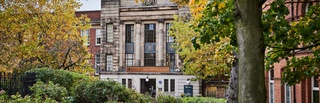 Exterior of the Wulfruna building, its name and the University of Wolverhampton logo visible on a large bronze sign above its double doors, photographed between a canopy of trees in St Peters gardens